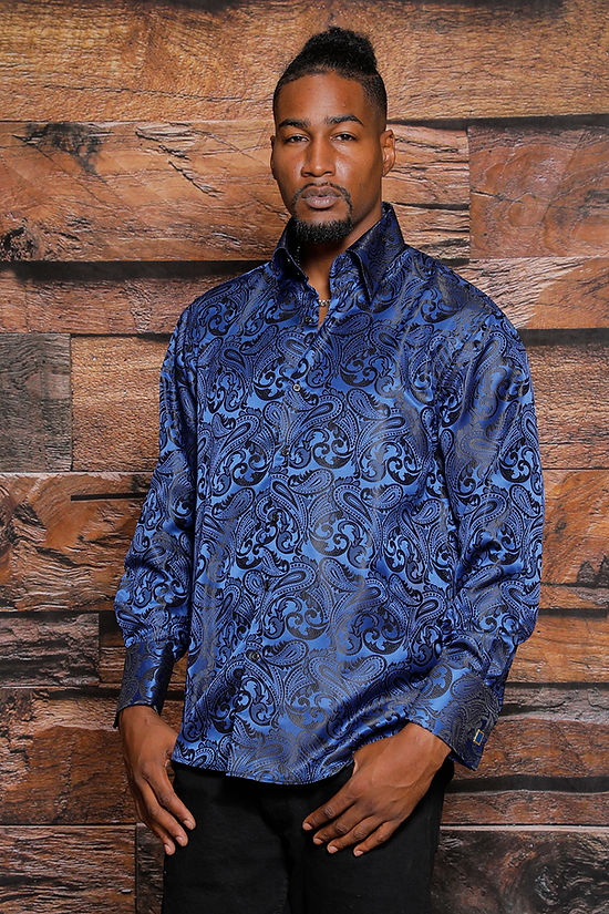 A man stands before a wooden wall wearing the Manzini MANZINI MZT-100 Paisley Long Sleeve Satin Navy Shirt w/ Links and black pants, looking at the camera. This stylish shirt by Manzini is perfect for formal occasions.