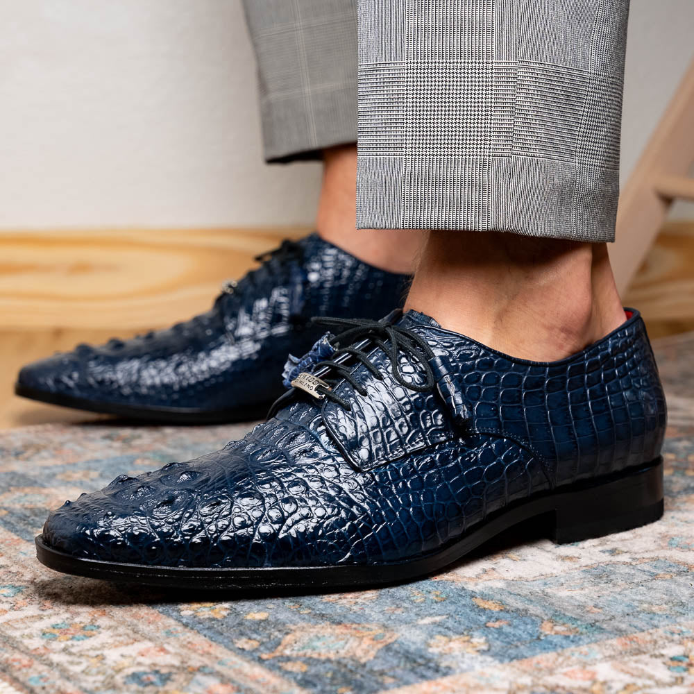 The Nilo Navy by Marco Di Milano features dark blue luxury footwear crafted from premium Nile crocodile skin with black laces and brown leather soles, displayed with one shoe propped on the other.
