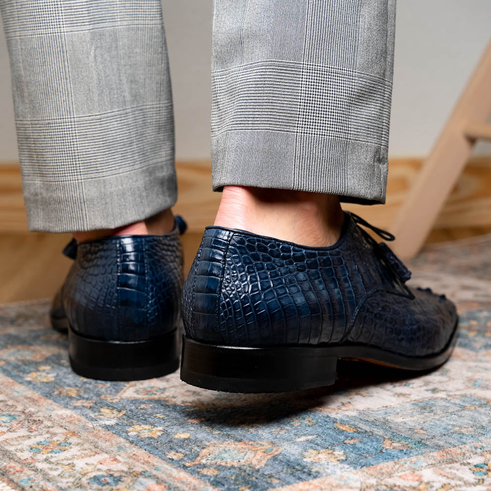 The Nilo Navy by Marco Di Milano features dark blue luxury footwear crafted from premium Nile crocodile skin with black laces and brown leather soles, displayed with one shoe propped on the other.