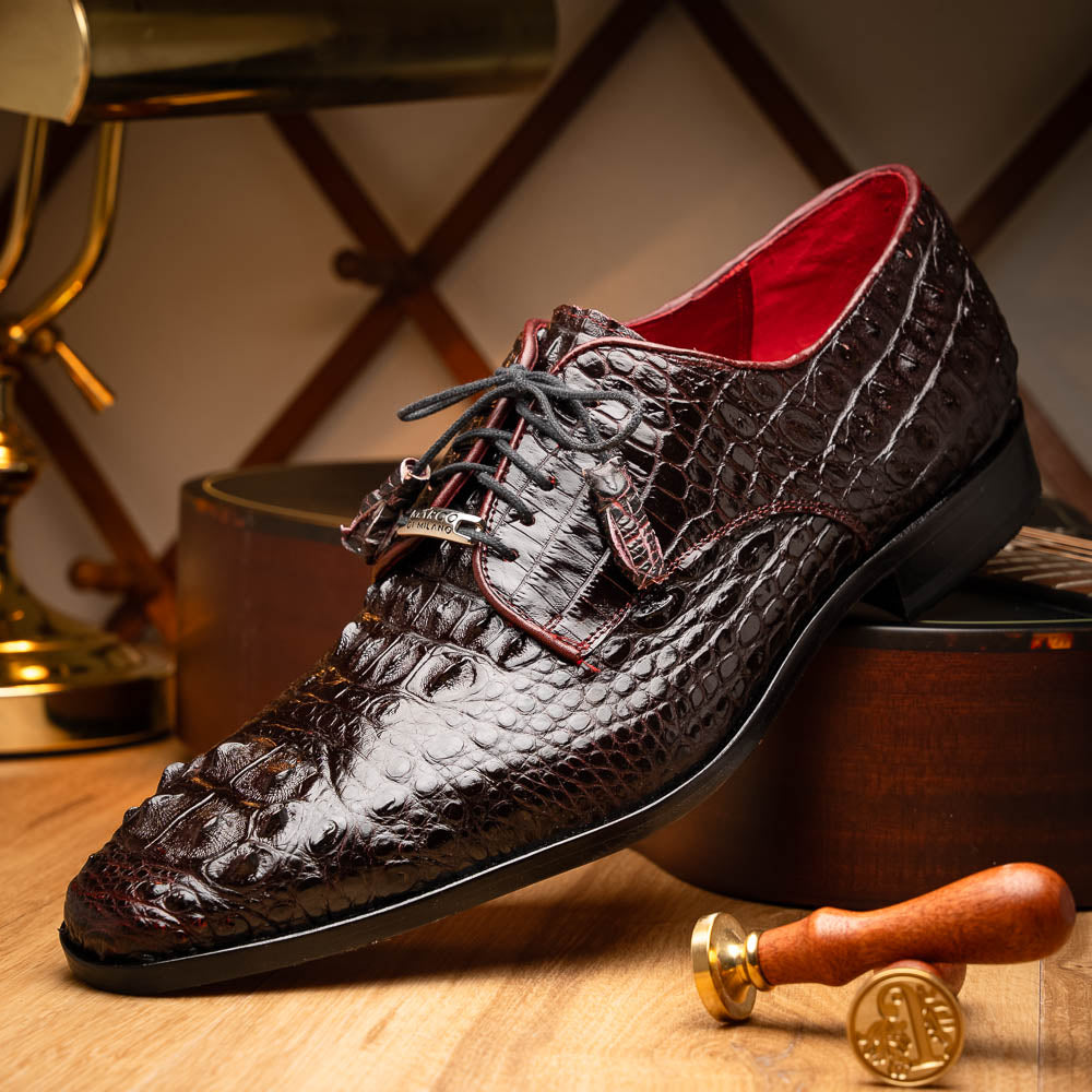 The Marco Di Milano Nilo Wine shoes are luxury dress footwear crafted from dark brown, textured Italian leather, featuring red interior lining and tan leather soles, with one shoe stylishly propped on the other.