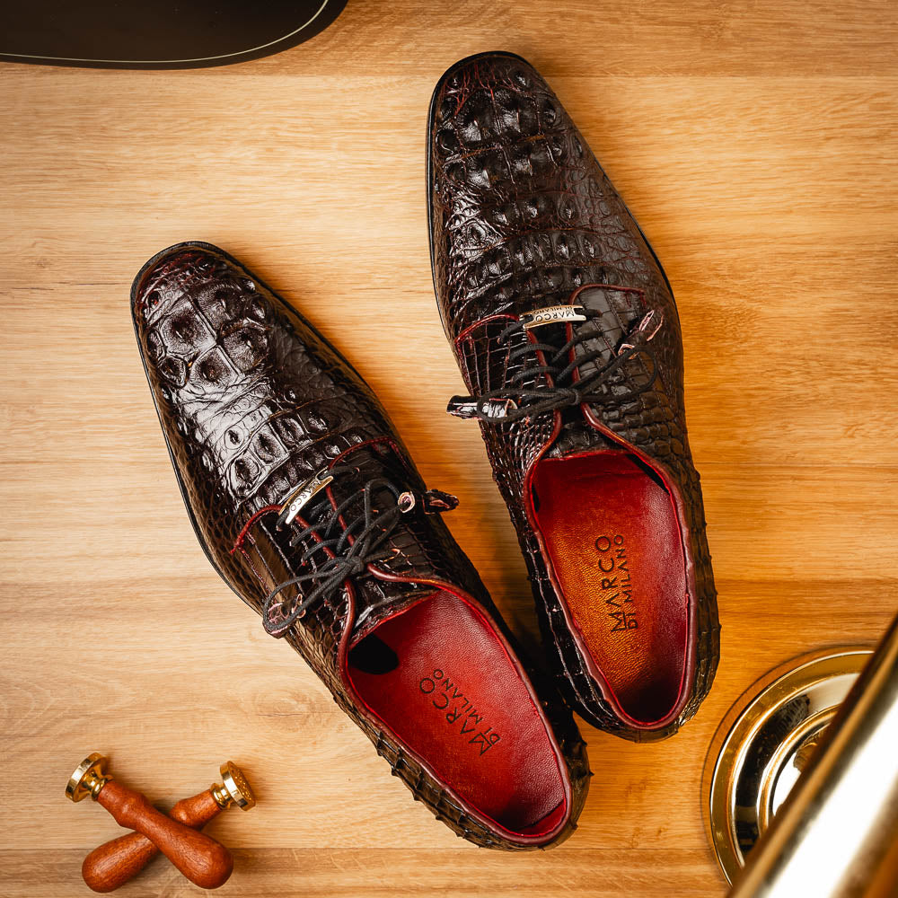 The Marco Di Milano Nilo Wine shoes are luxury dress footwear crafted from dark brown, textured Italian leather, featuring red interior lining and tan leather soles, with one shoe stylishly propped on the other.
