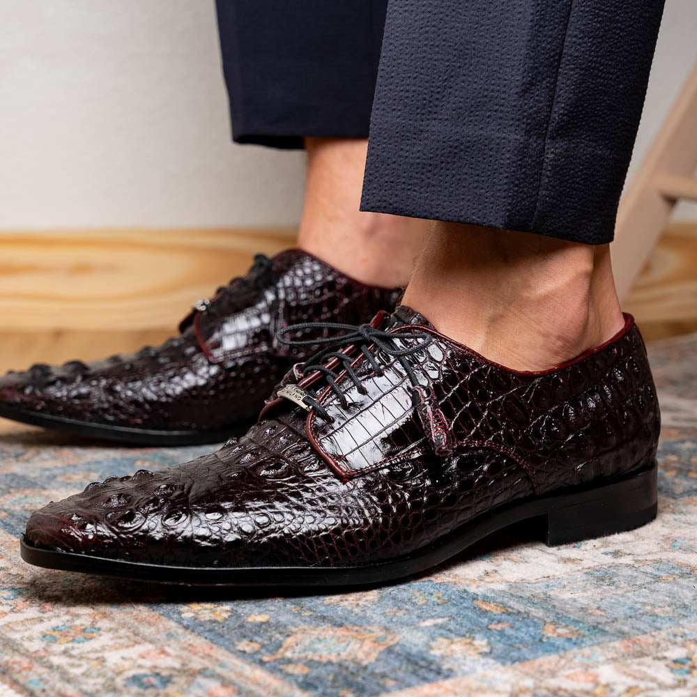 The Marco Di Milano Nilo Wine shoes are luxury dress footwear crafted from dark brown, textured Italian leather, featuring red interior lining and tan leather soles, with one shoe stylishly propped on the other.