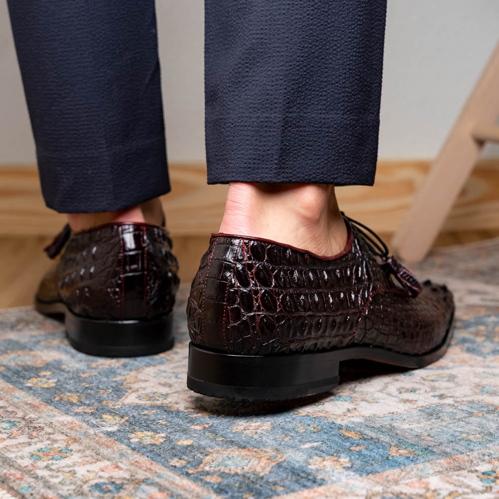The Marco Di Milano Nilo Wine shoes are luxury dress footwear crafted from dark brown, textured Italian leather, featuring red interior lining and tan leather soles, with one shoe stylishly propped on the other.