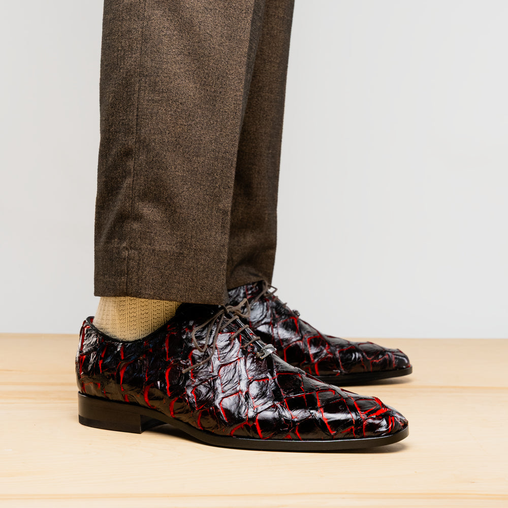 A pair of Marco Di Milano Olivieri Blackcherry oxfords—one upright, one on its side—displays the brand name on the sole, offering luxury men’s footwear with a strikingly exotic black and red pirarucu finish.