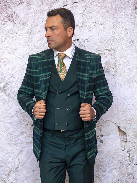A man wears the Manzini PARIS-1 Men's 3-Piece Tailored Fit Plaid Business Suit in forest green with a patterned tie, standing against a textured light wall, looking to the side while holding his jacket.
