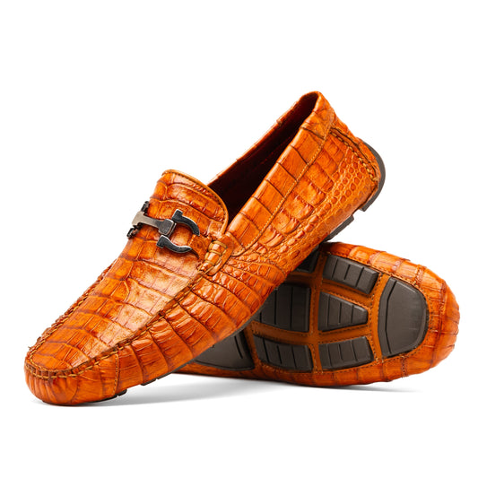 Marco Di Milano’s Parma Brandy loafers feature an orange crocodile pattern, black metal buckles, and hand-painted finishes—one shoe stands upright while the other lies on its side to reveal the detailed sole.