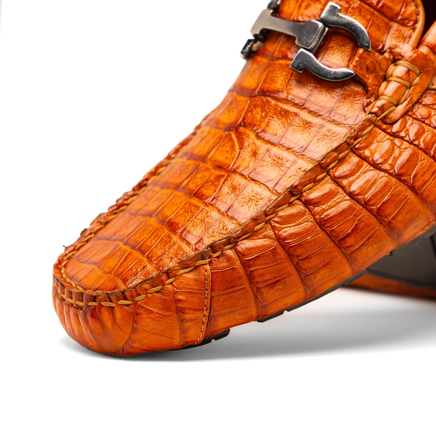 Marco Di Milano’s Parma Brandy loafers feature an orange crocodile pattern, black metal buckles, and hand-painted finishes—one shoe stands upright while the other lies on its side to reveal the detailed sole.