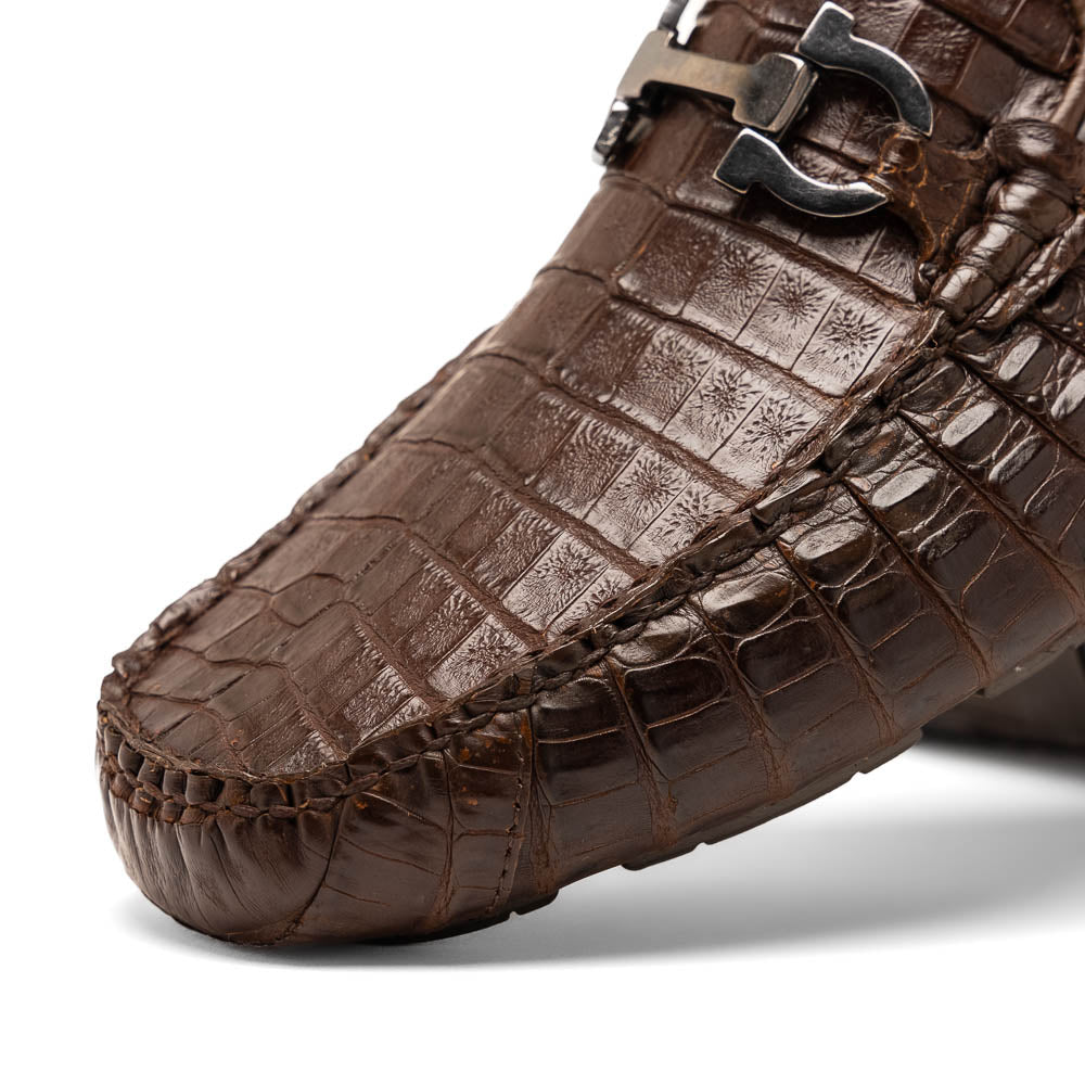 The Marco Di Milano Parma Brown features a pair of brown caiman belly loafers with black metal buckles—one upright, the other on its side showing the sole—a luxurious footwear choice.