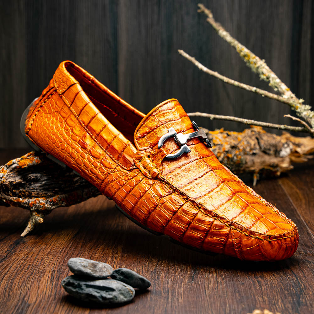 Marco Di Milano’s Parma Brandy loafers feature an orange crocodile pattern, black metal buckles, and hand-painted finishes—one shoe stands upright while the other lies on its side to reveal the detailed sole.
