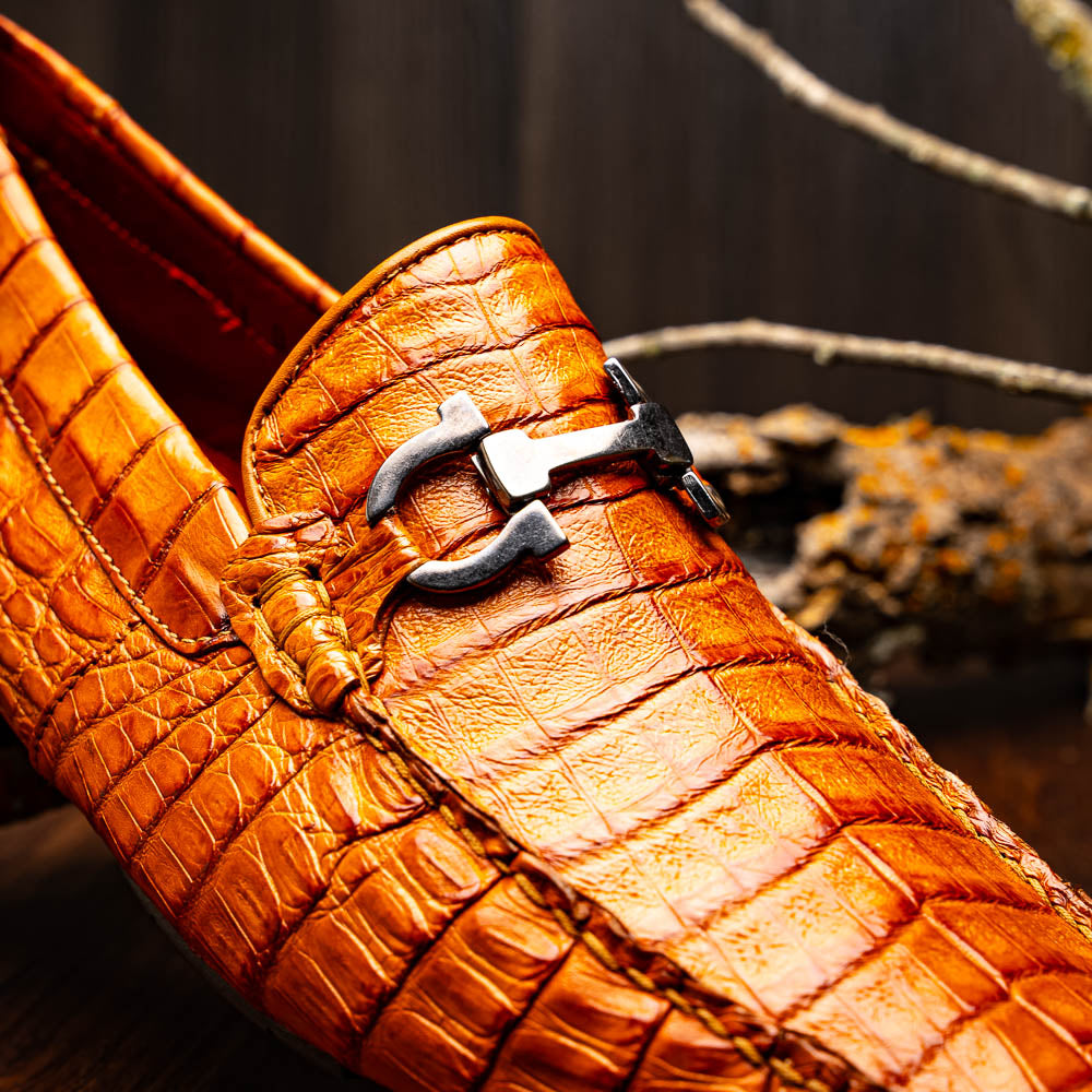 Marco Di Milano’s Parma Brandy loafers feature an orange crocodile pattern, black metal buckles, and hand-painted finishes—one shoe stands upright while the other lies on its side to reveal the detailed sole.