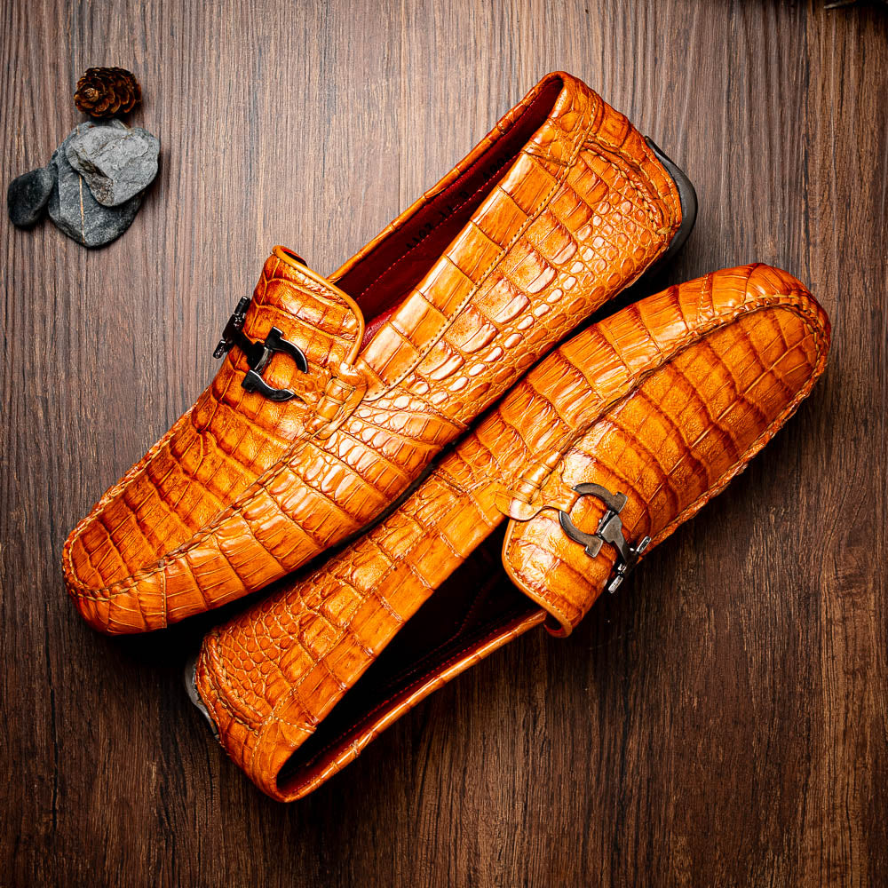 Marco Di Milano’s Parma Brandy loafers feature an orange crocodile pattern, black metal buckles, and hand-painted finishes—one shoe stands upright while the other lies on its side to reveal the detailed sole.