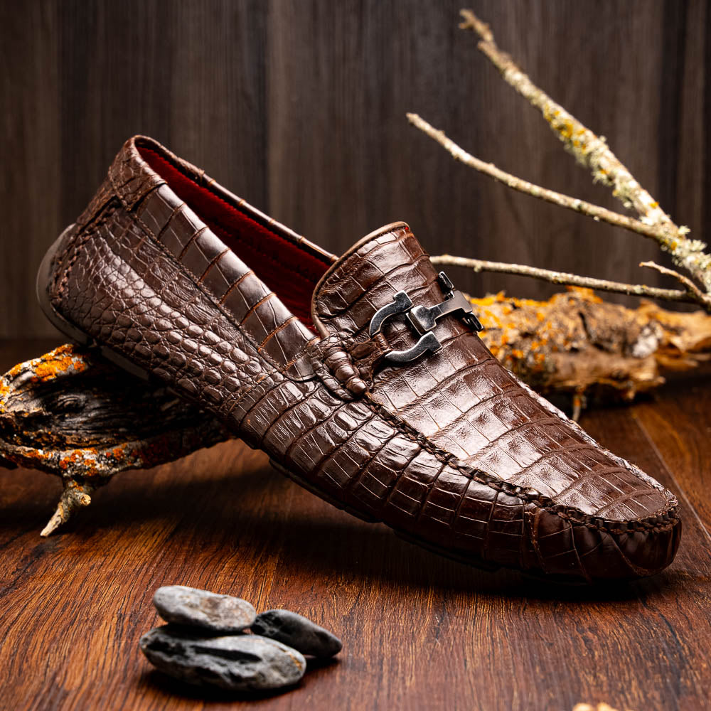 The Marco Di Milano Parma Brown features a pair of brown caiman belly loafers with black metal buckles—one upright, the other on its side showing the sole—a luxurious footwear choice.