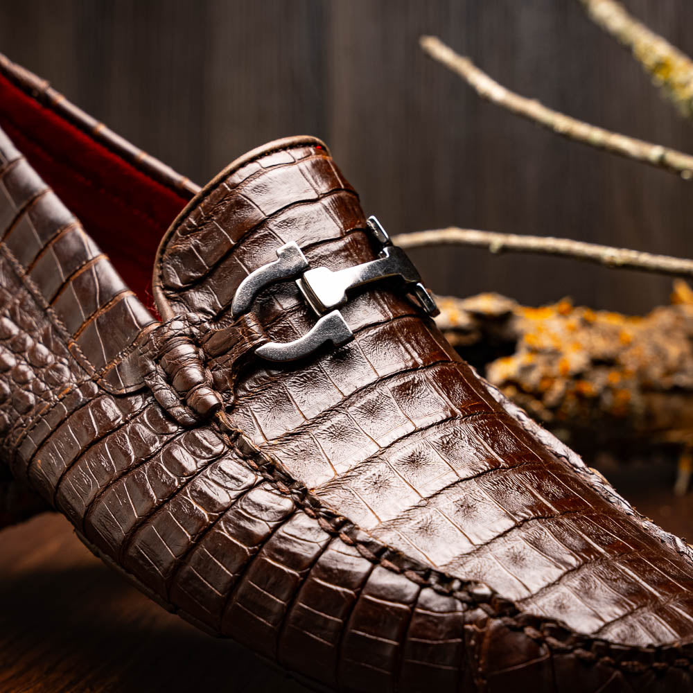 The Marco Di Milano Parma Brown features a pair of brown caiman belly loafers with black metal buckles—one upright, the other on its side showing the sole—a luxurious footwear choice.