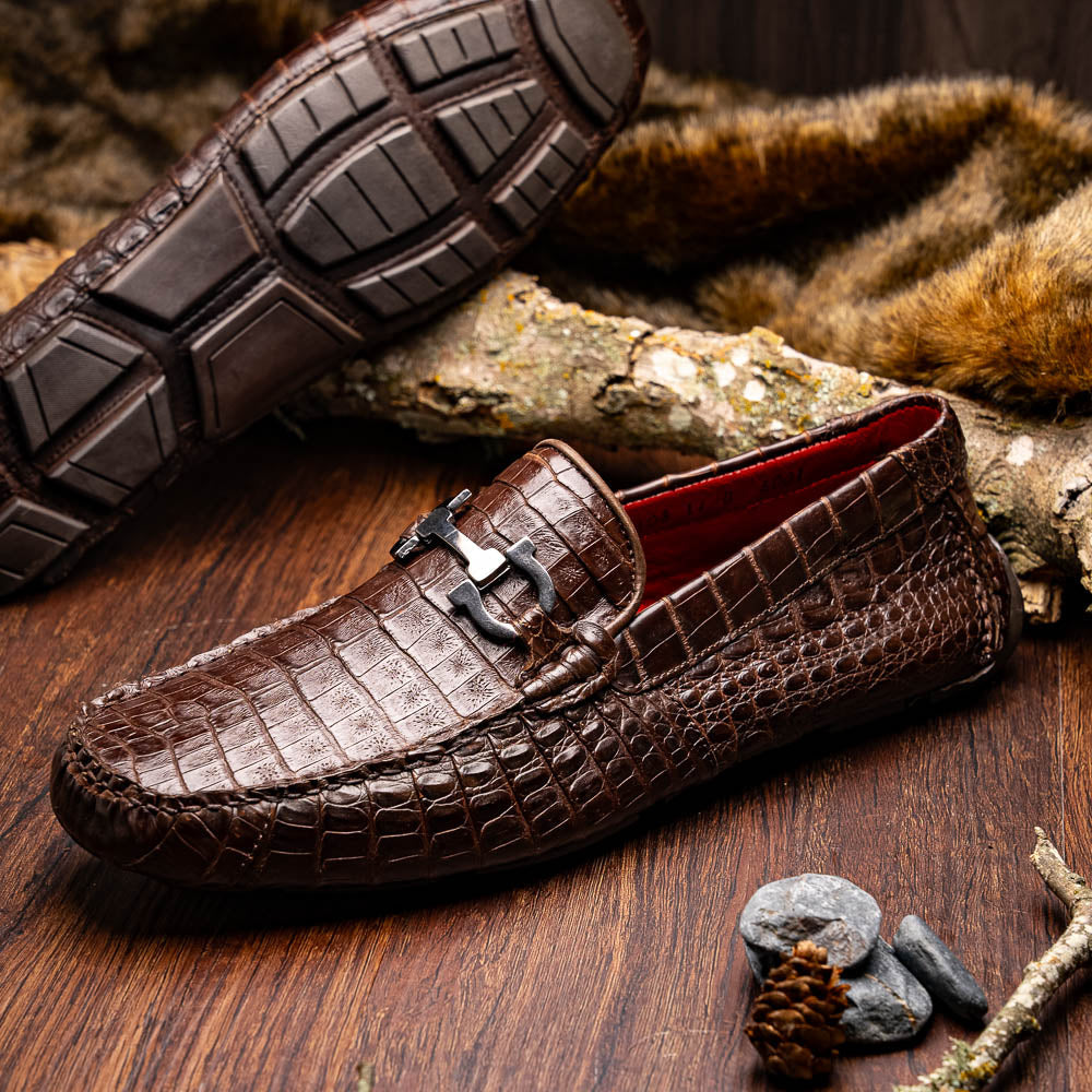 The Marco Di Milano Parma Brown features a pair of brown caiman belly loafers with black metal buckles—one upright, the other on its side showing the sole—a luxurious footwear choice.