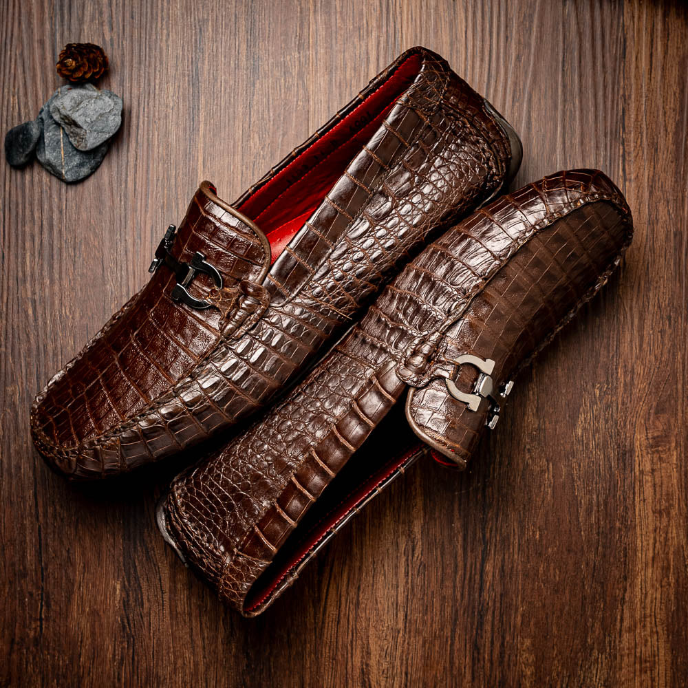 The Marco Di Milano Parma Brown features a pair of brown caiman belly loafers with black metal buckles—one upright, the other on its side showing the sole—a luxurious footwear choice.