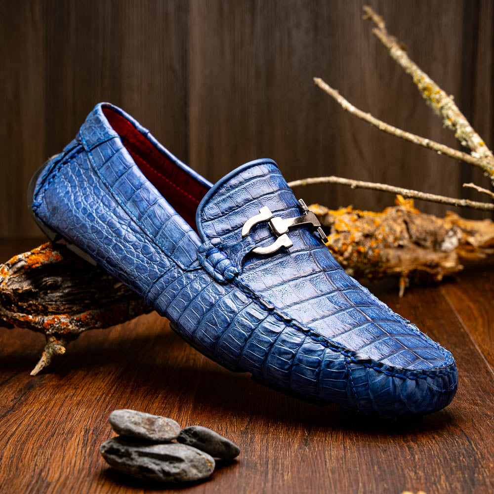 The Marco Di Milano Parma Blue Jeans loafers feature genuine blue caiman leather with a crocodile pattern, metal hardware accents, and rubber soles—ideal luxury footwear, displayed with one shoe upright and the other on its side.