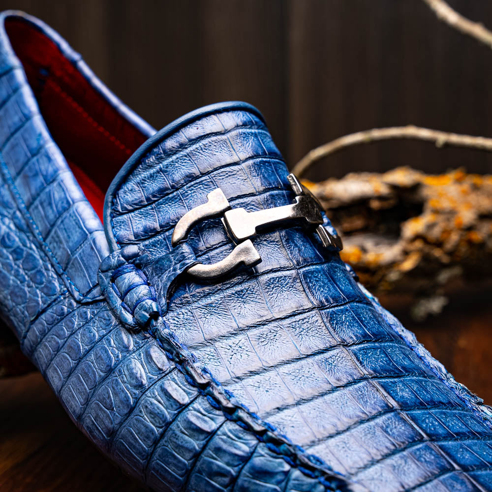 The Marco Di Milano Parma Blue Jeans loafers feature genuine blue caiman leather with a crocodile pattern, metal hardware accents, and rubber soles—ideal luxury footwear, displayed with one shoe upright and the other on its side.