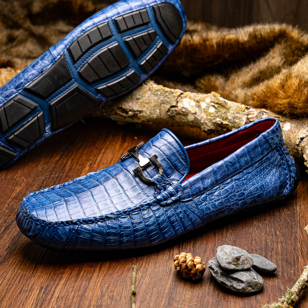 The Marco Di Milano Parma Blue Jeans loafers feature genuine blue caiman leather with a crocodile pattern, metal hardware accents, and rubber soles—ideal luxury footwear, displayed with one shoe upright and the other on its side.