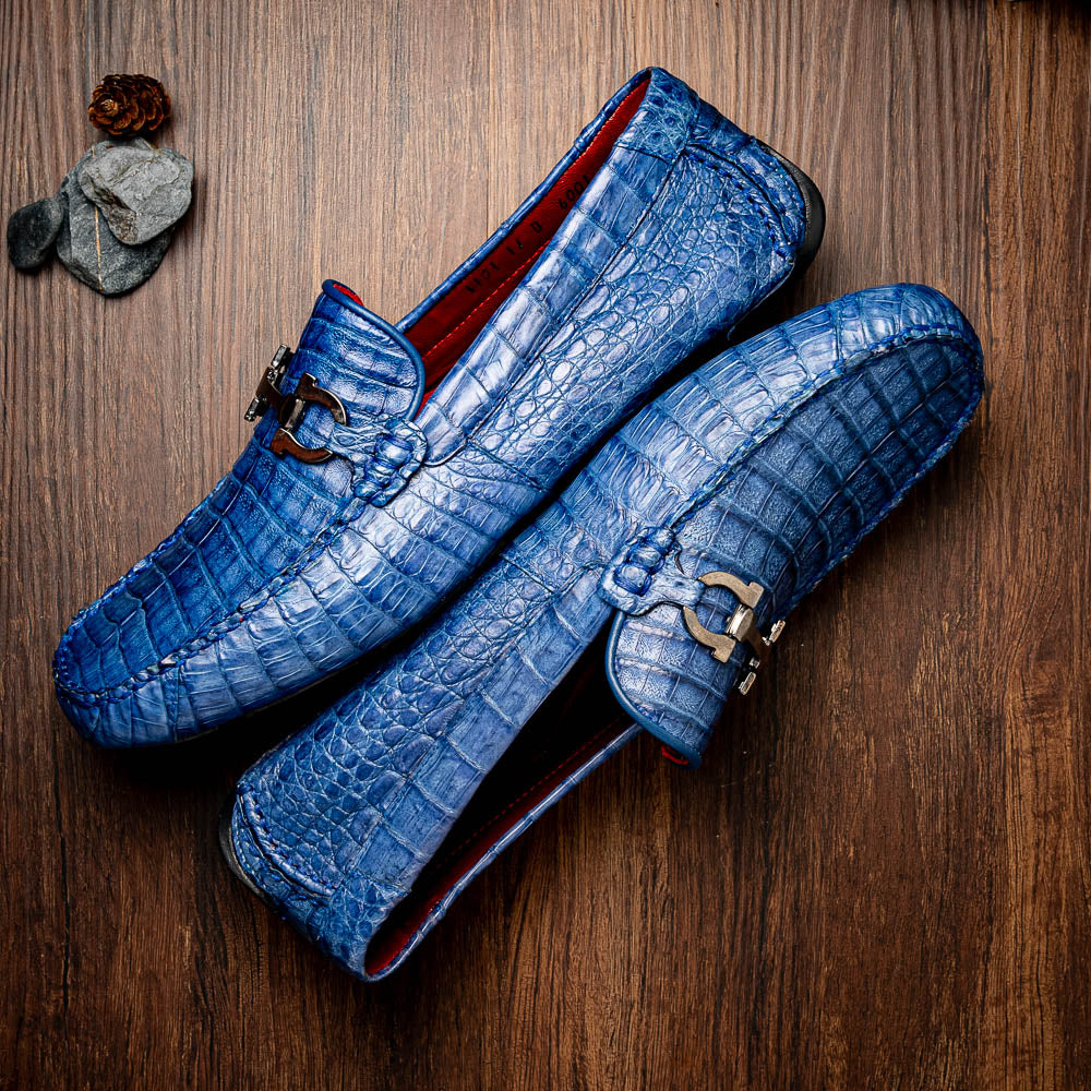 The Marco Di Milano Parma Blue Jeans loafers feature genuine blue caiman leather with a crocodile pattern, metal hardware accents, and rubber soles—ideal luxury footwear, displayed with one shoe upright and the other on its side.