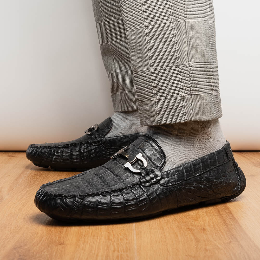The Marco Di Milano Parma Black is a luxury caiman belly loafer featuring black crocodile-textured leather, metal buckle accents, and rubber soles, with one men’s driving shoe upright and the other leaning against it.