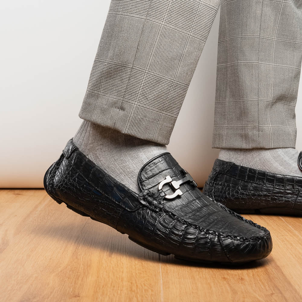 The Marco Di Milano Parma Black is a luxury caiman belly loafer featuring black crocodile-textured leather, metal buckle accents, and rubber soles, with one men’s driving shoe upright and the other leaning against it.