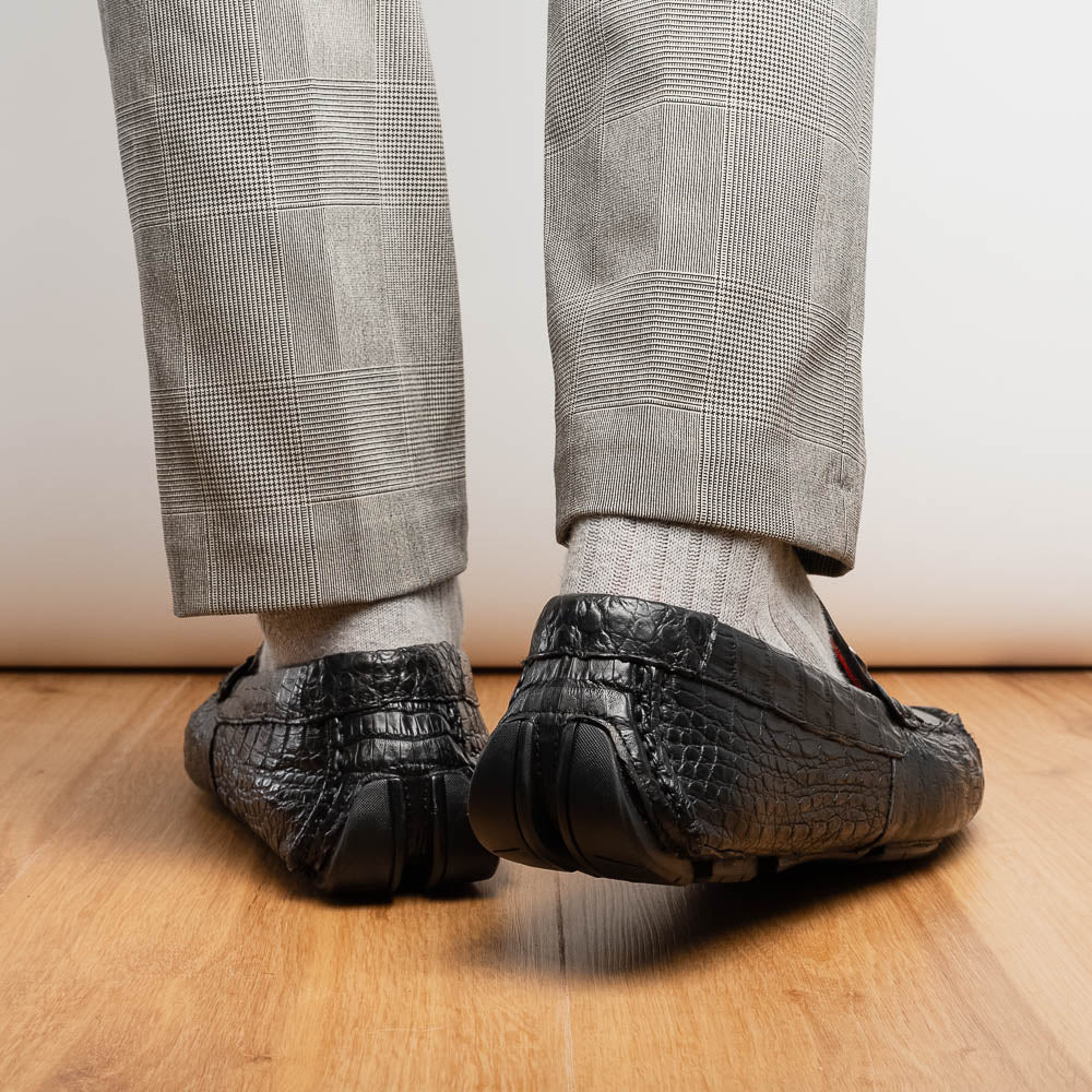The Marco Di Milano Parma Black is a luxury caiman belly loafer featuring black crocodile-textured leather, metal buckle accents, and rubber soles, with one men’s driving shoe upright and the other leaning against it.