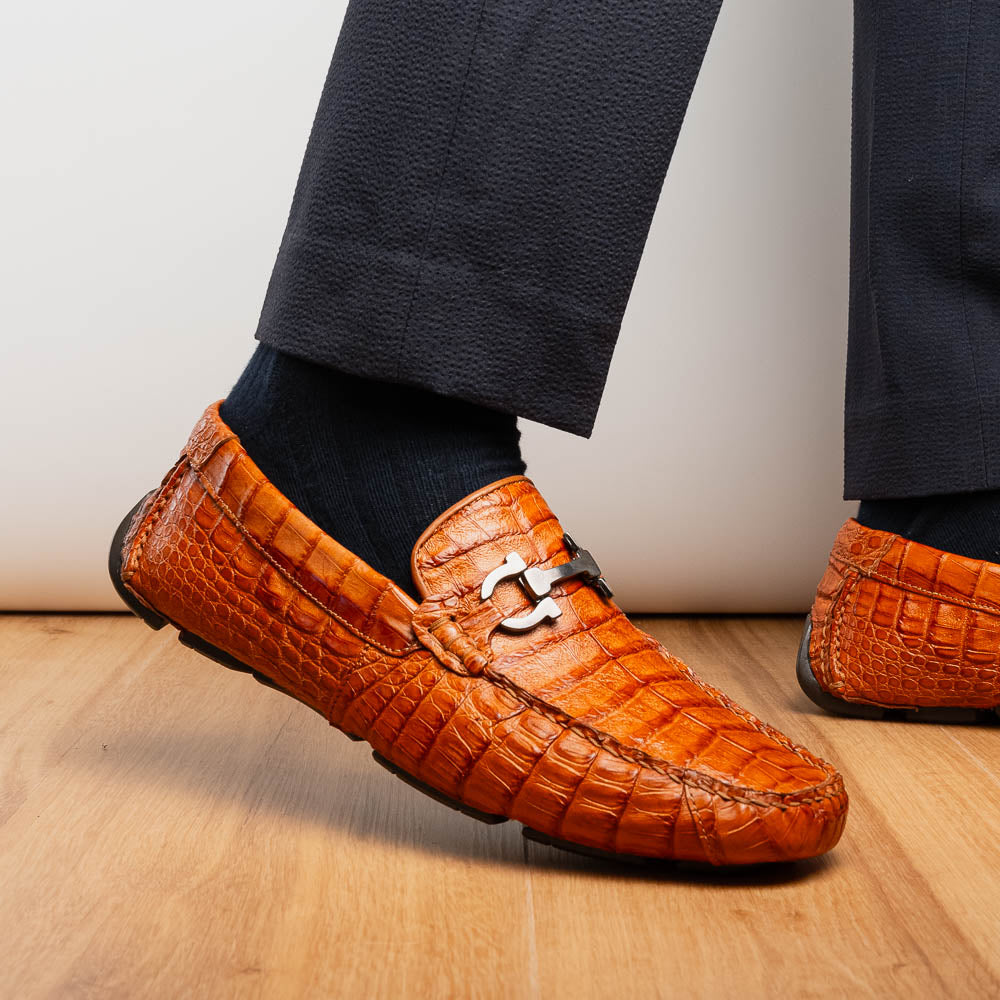 Marco Di Milano’s Parma Brandy loafers feature an orange crocodile pattern, black metal buckles, and hand-painted finishes—one shoe stands upright while the other lies on its side to reveal the detailed sole.