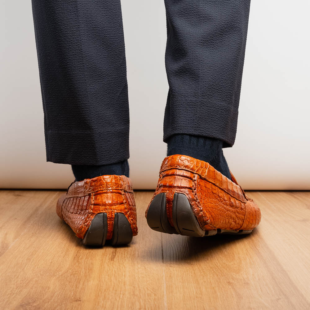 Marco Di Milano’s Parma Brandy loafers feature an orange crocodile pattern, black metal buckles, and hand-painted finishes—one shoe stands upright while the other lies on its side to reveal the detailed sole.
