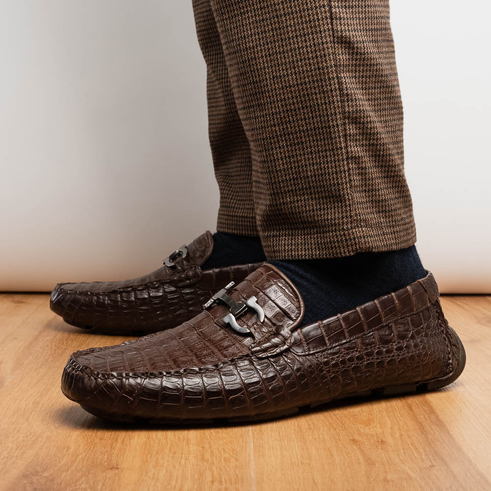 The Marco Di Milano Parma Brown features a pair of brown caiman belly loafers with black metal buckles—one upright, the other on its side showing the sole—a luxurious footwear choice.