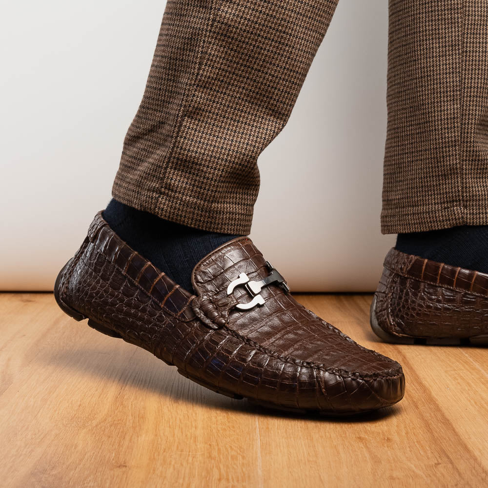 The Marco Di Milano Parma Brown features a pair of brown caiman belly loafers with black metal buckles—one upright, the other on its side showing the sole—a luxurious footwear choice.