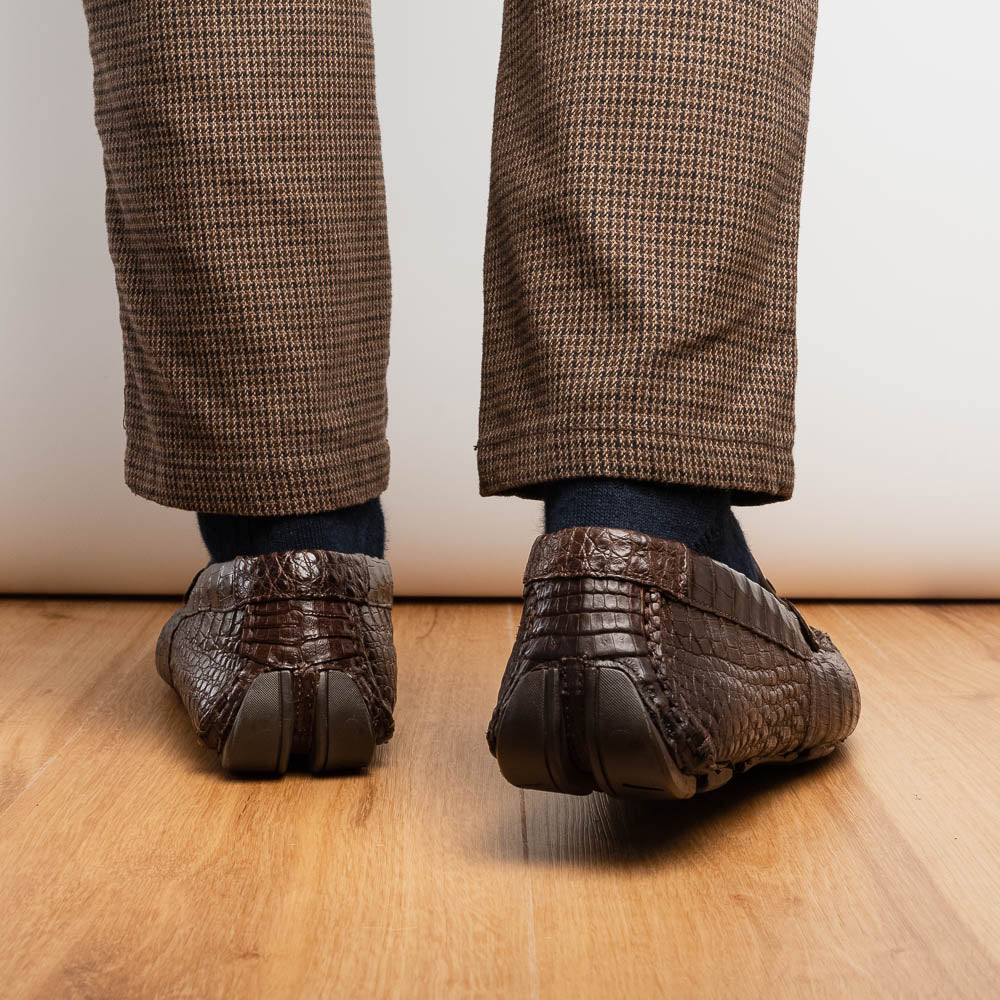 The Marco Di Milano Parma Brown features a pair of brown caiman belly loafers with black metal buckles—one upright, the other on its side showing the sole—a luxurious footwear choice.