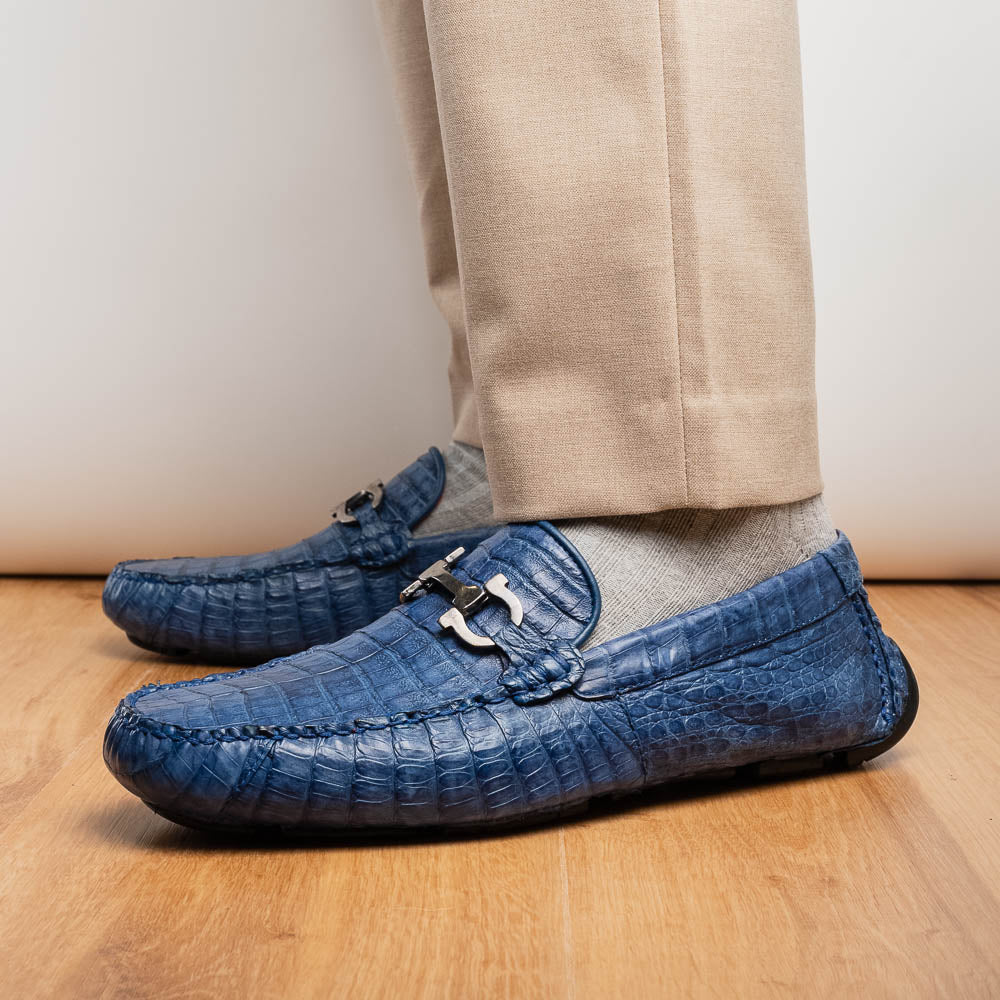 The Marco Di Milano Parma Blue Jeans loafers feature genuine blue caiman leather with a crocodile pattern, metal hardware accents, and rubber soles—ideal luxury footwear, displayed with one shoe upright and the other on its side.