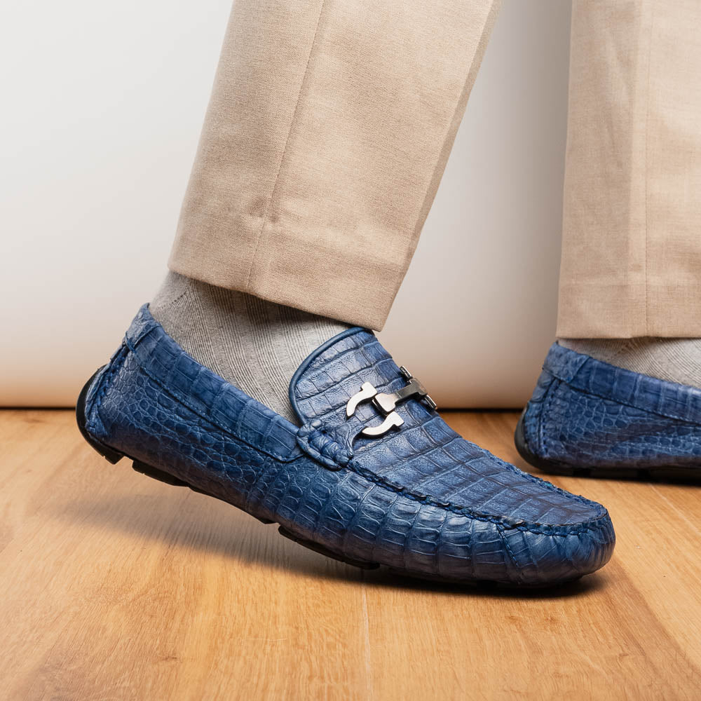 The Marco Di Milano Parma Blue Jeans loafers feature genuine blue caiman leather with a crocodile pattern, metal hardware accents, and rubber soles—ideal luxury footwear, displayed with one shoe upright and the other on its side.