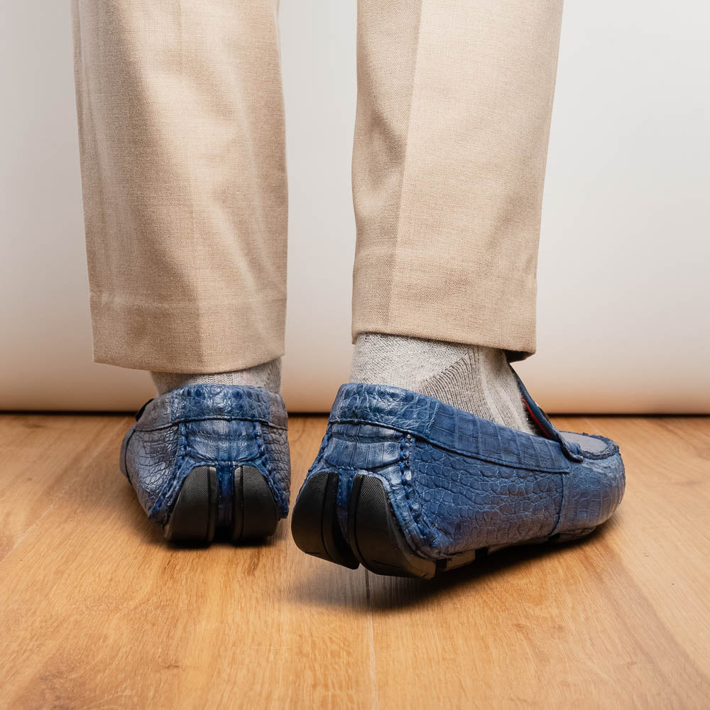 The Marco Di Milano Parma Blue Jeans loafers feature genuine blue caiman leather with a crocodile pattern, metal hardware accents, and rubber soles—ideal luxury footwear, displayed with one shoe upright and the other on its side.