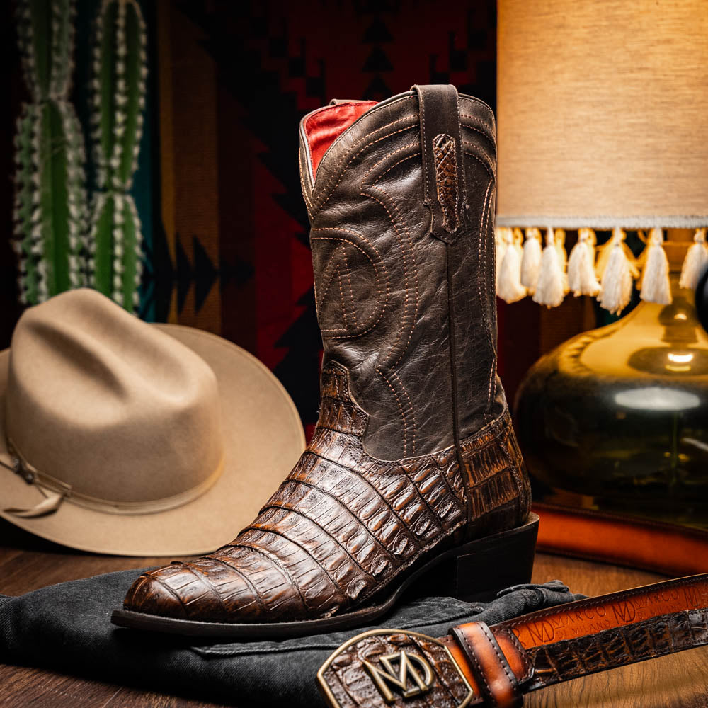 A pair of Marco Di Milano Phoenix Aged Brown men's boots, one upright and one showing the sole, crafted from genuine caiman belly skin with a crocodile texture and detailed stitching—ideal for any Western collection.