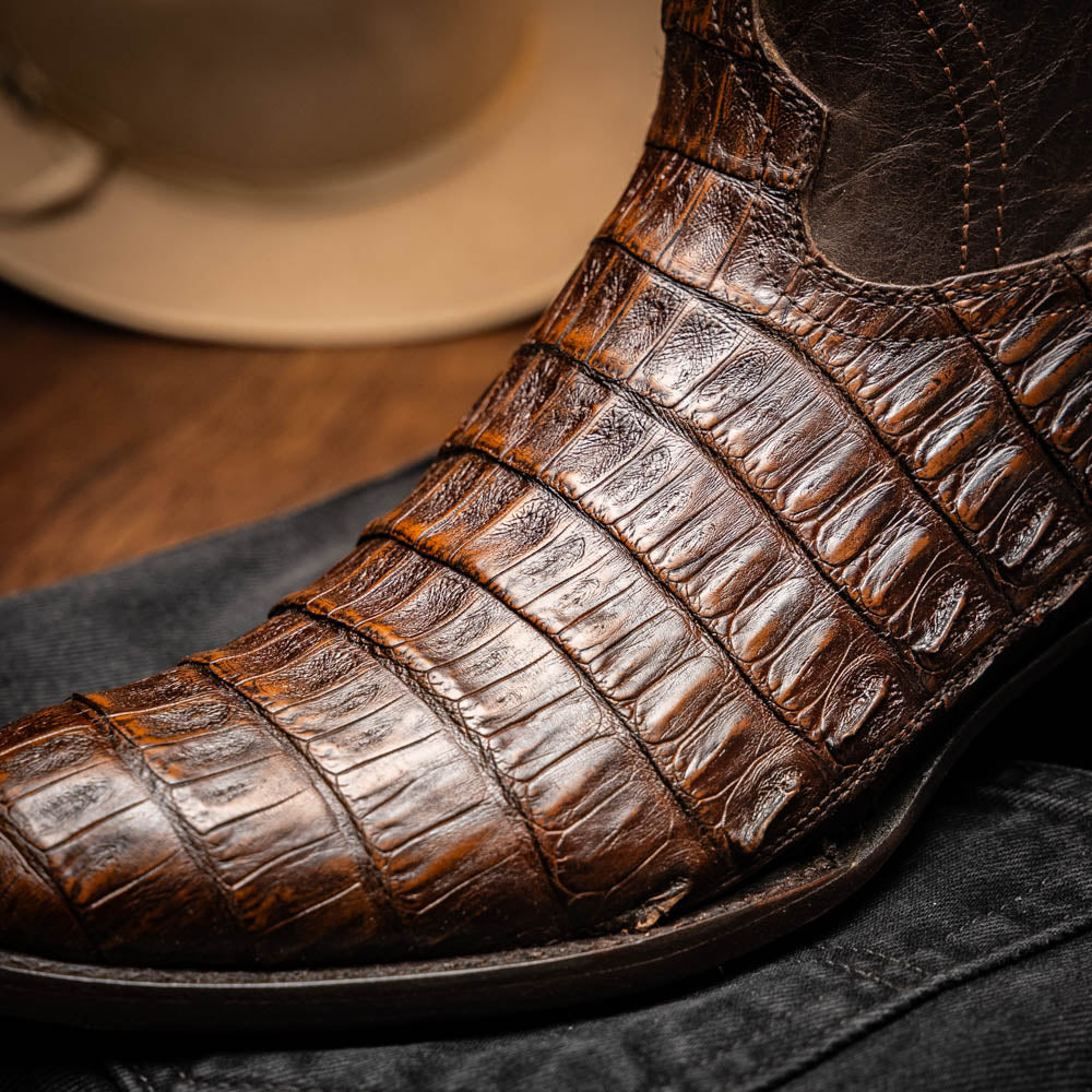 A pair of Marco Di Milano Phoenix Aged Brown men's boots, one upright and one showing the sole, crafted from genuine caiman belly skin with a crocodile texture and detailed stitching—ideal for any Western collection.