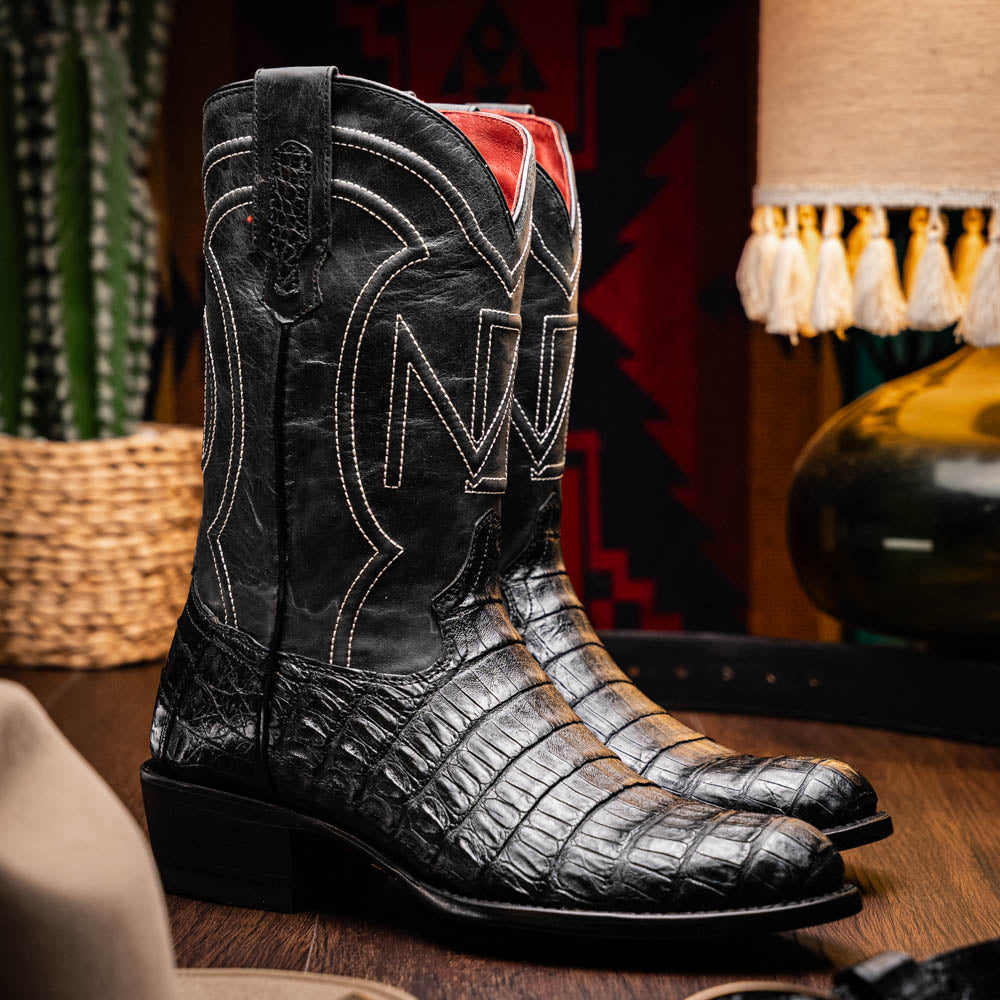 A pair of Phoenix Black Western cowboy boots by Marco Di Milano, one upright and one showing the sole, featuring a textured pattern and the brand name on the sole.