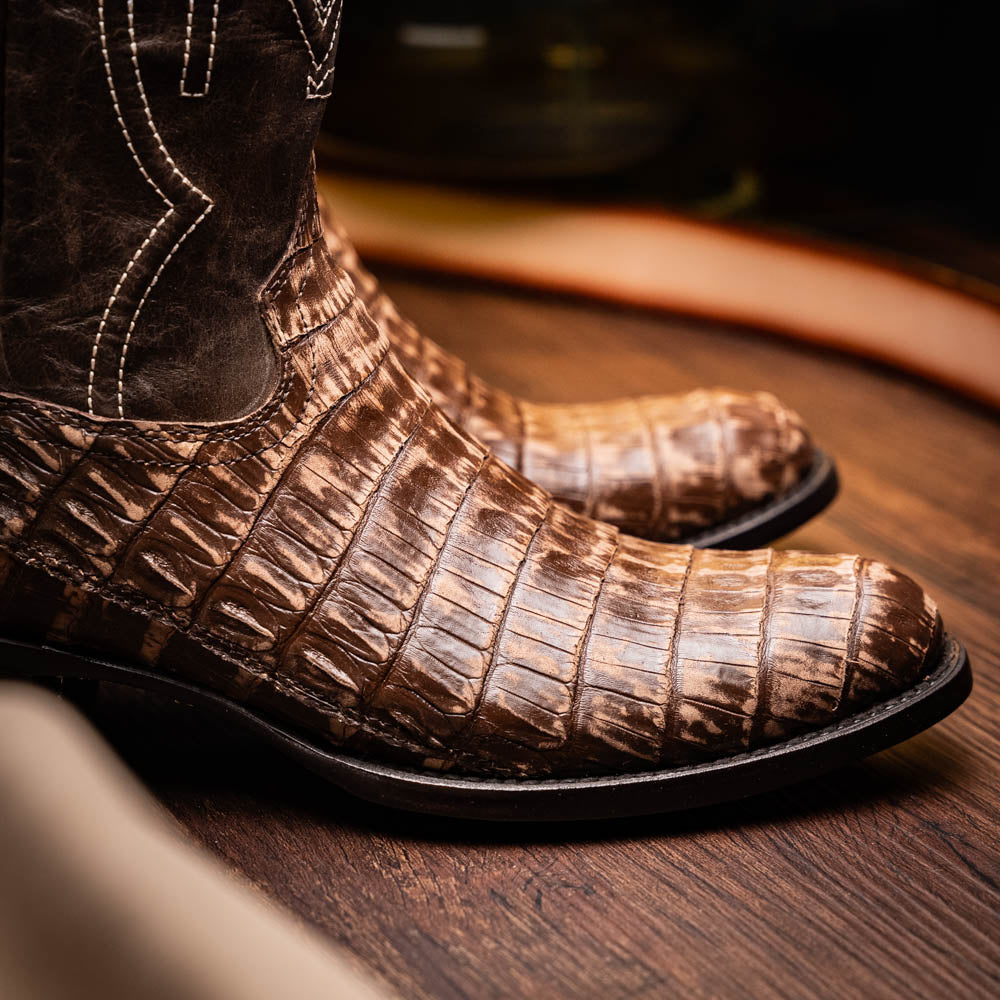 The Phoenix Stone Brown by Marco Di Milano are brown Western cowboy boots featuring crocodile-patterned textured leather, decorative stitching, and branded soles.