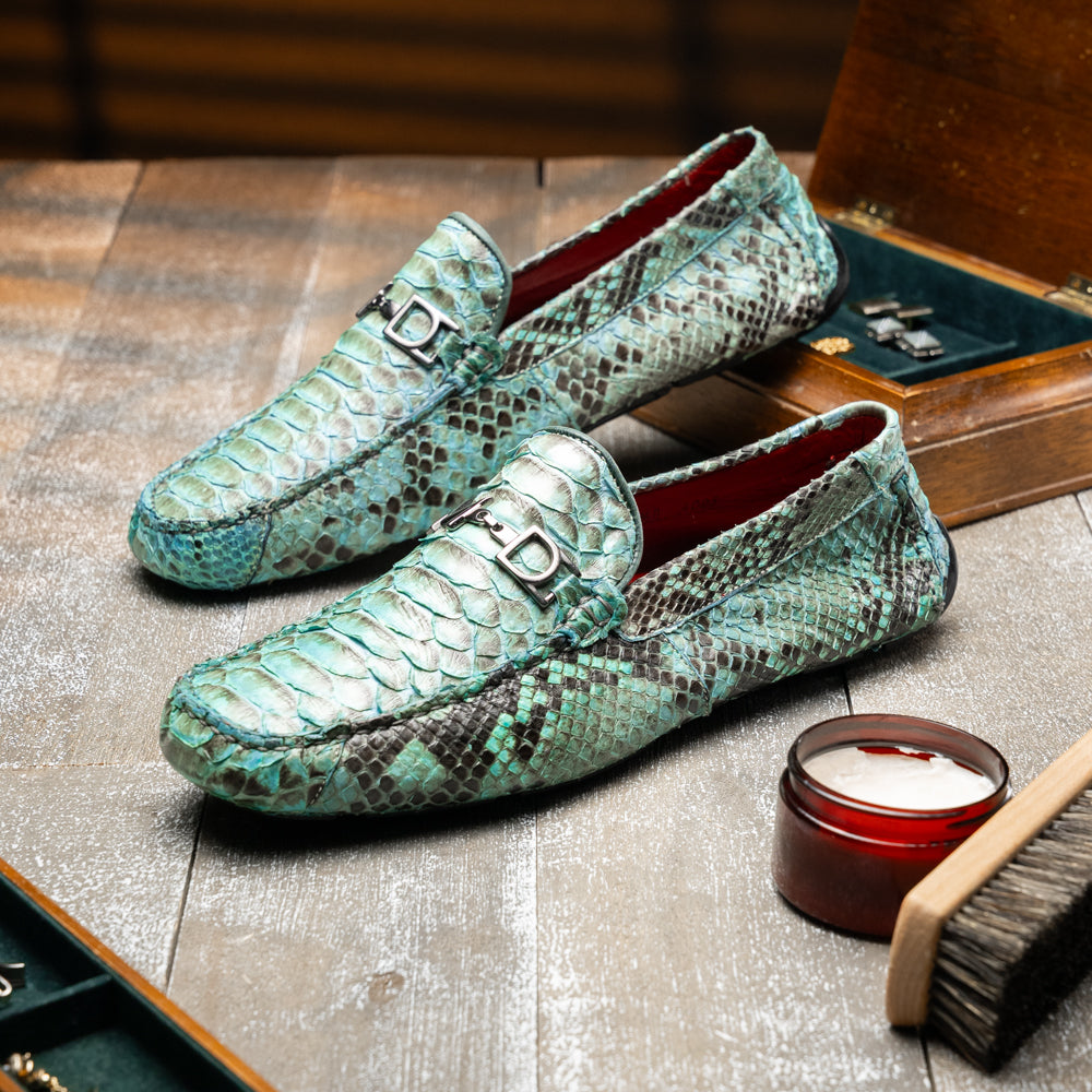 Introducing the Pia Aqua by Marco Di Milano—a pair of luxury men’s loafers handcrafted from striking green python skin. Featuring black rubber soles and silver buckle accents, one shoe is elegantly displayed on its side to highlight the distinctive design of this opulent driver shoe.