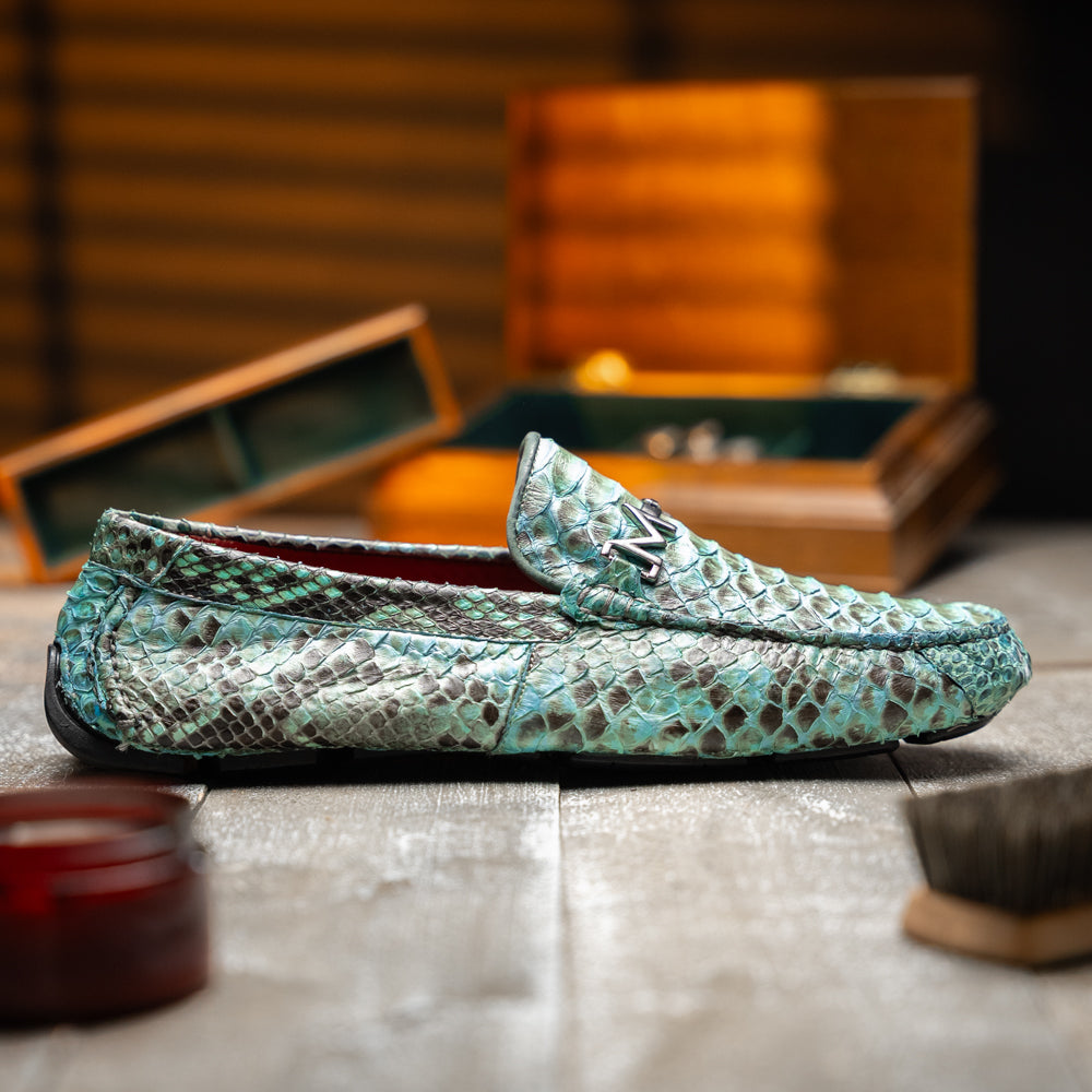 Introducing the Pia Aqua by Marco Di Milano—a pair of luxury men’s loafers handcrafted from striking green python skin. Featuring black rubber soles and silver buckle accents, one shoe is elegantly displayed on its side to highlight the distinctive design of this opulent driver shoe.