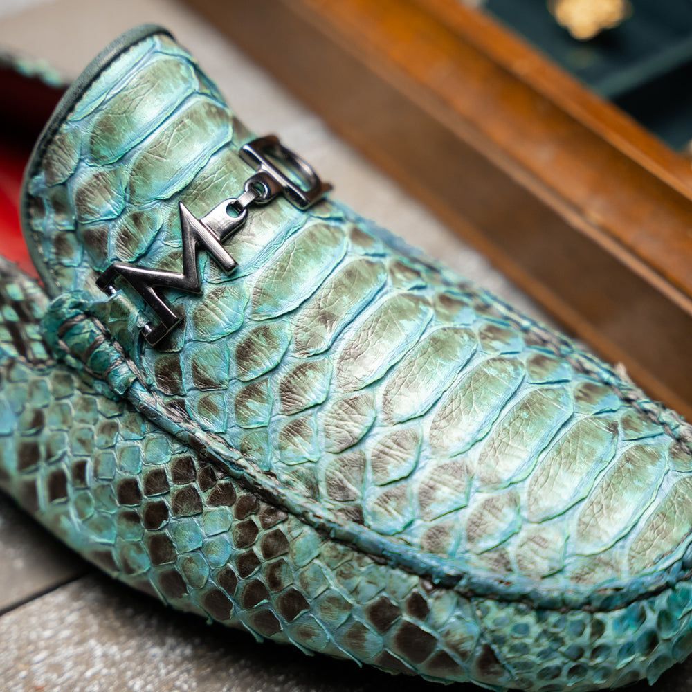 Introducing the Pia Aqua by Marco Di Milano—a pair of luxury men’s loafers handcrafted from striking green python skin. Featuring black rubber soles and silver buckle accents, one shoe is elegantly displayed on its side to highlight the distinctive design of this opulent driver shoe.