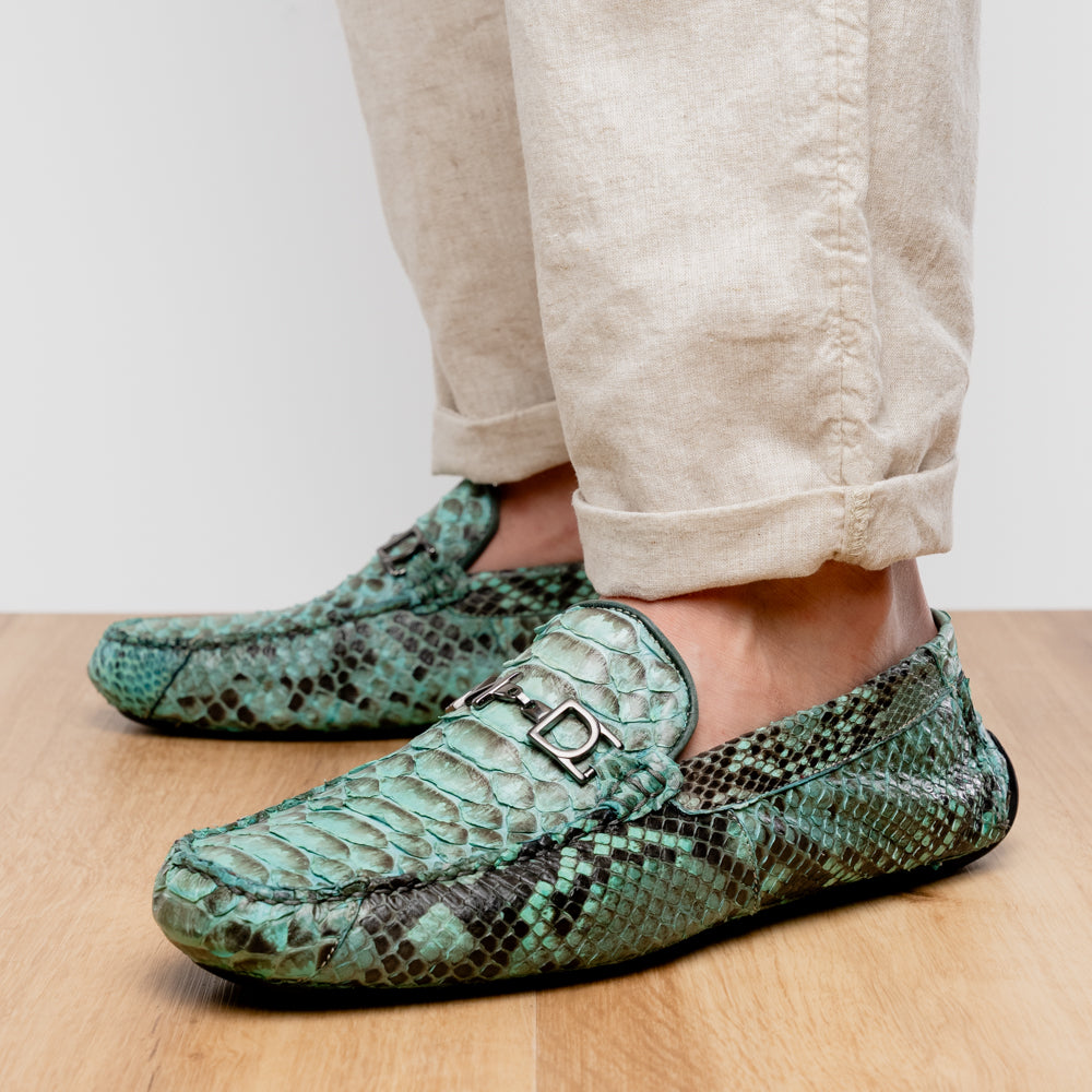 Introducing the Pia Aqua by Marco Di Milano—a pair of luxury men’s loafers handcrafted from striking green python skin. Featuring black rubber soles and silver buckle accents, one shoe is elegantly displayed on its side to highlight the distinctive design of this opulent driver shoe.