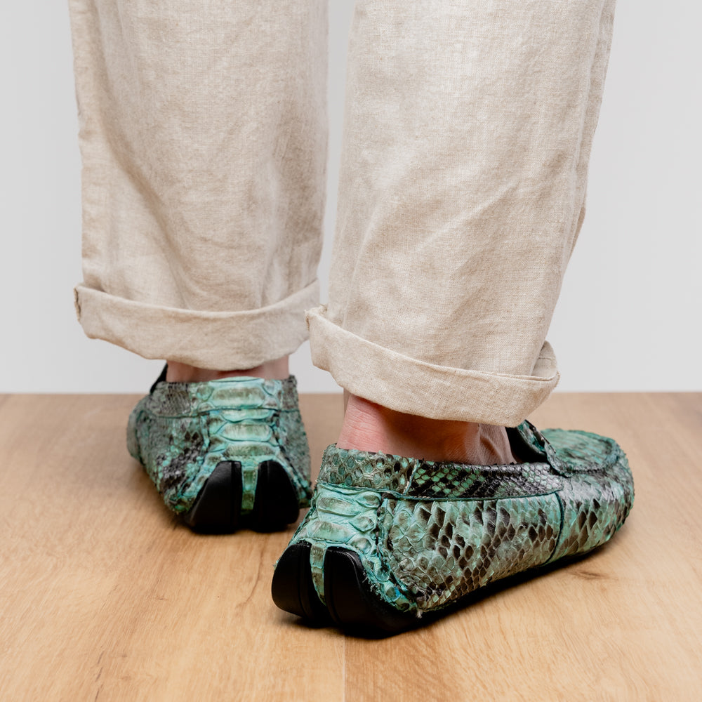 Introducing the Pia Aqua by Marco Di Milano—a pair of luxury men’s loafers handcrafted from striking green python skin. Featuring black rubber soles and silver buckle accents, one shoe is elegantly displayed on its side to highlight the distinctive design of this opulent driver shoe.