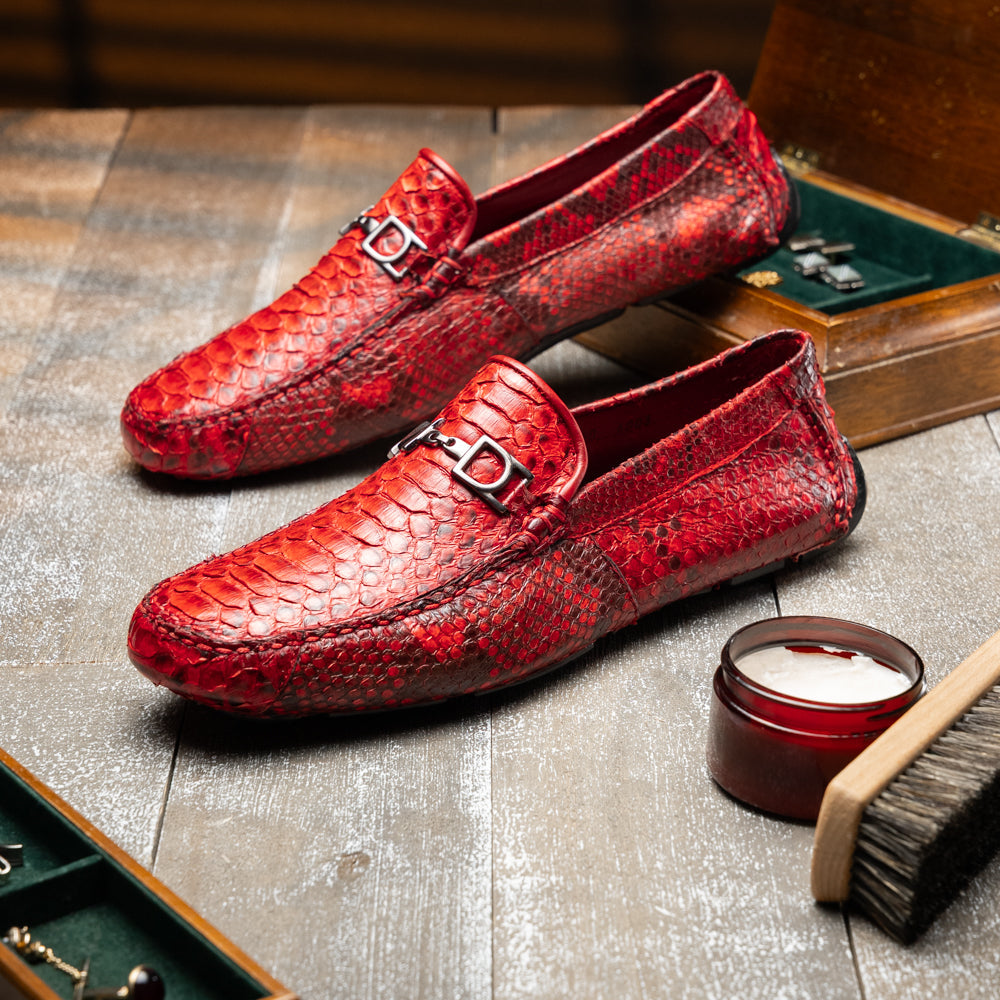 A pair of Pia Red loafers by Marco Di Milano—crafted as luxury men’s driver shoes in a striking red snakeskin pattern, featuring black rubber soles and silver buckle detailing, with one shoe upright and the other displayed on its side—showcase the allure of exotic python-inspired style in a distinguished design.