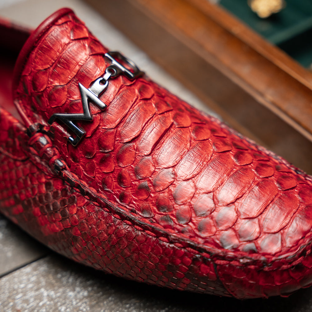 A pair of Pia Red loafers by Marco Di Milano—crafted as luxury men’s driver shoes in a striking red snakeskin pattern, featuring black rubber soles and silver buckle detailing, with one shoe upright and the other displayed on its side—showcase the allure of exotic python-inspired style in a distinguished design.