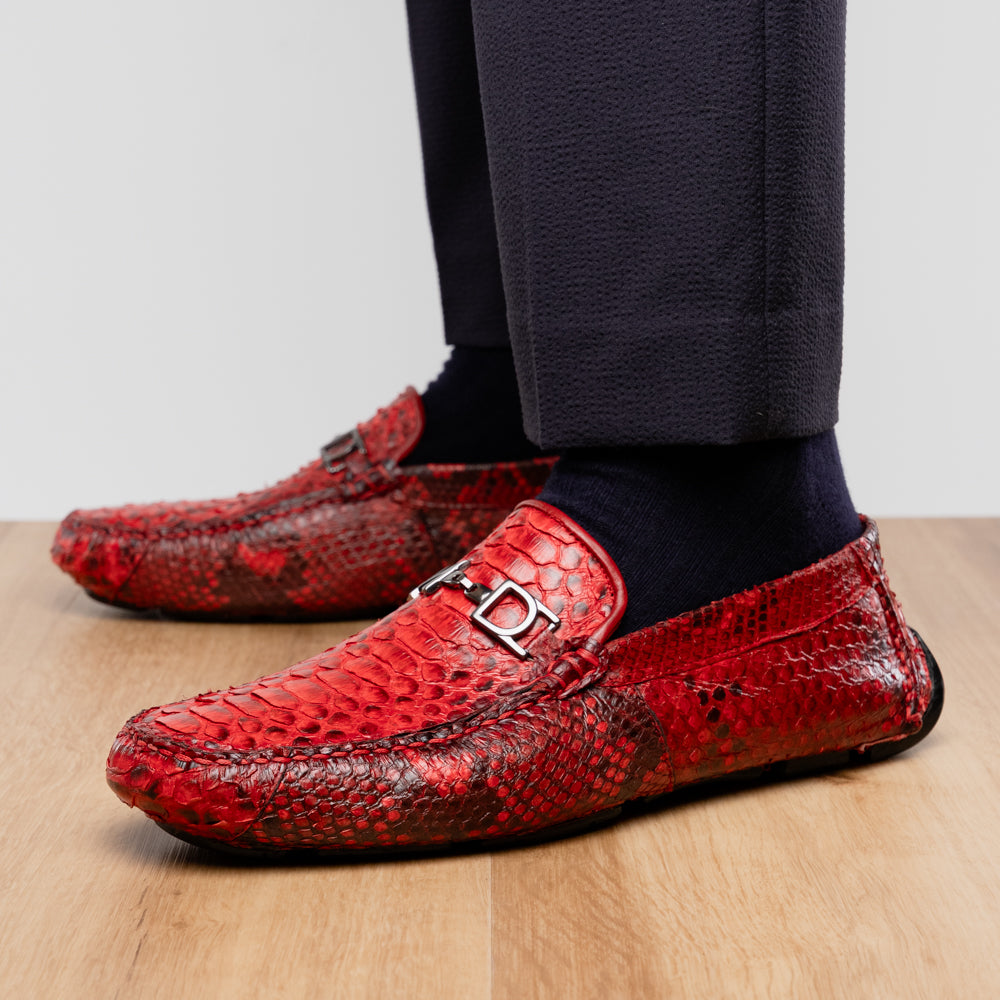 A pair of Pia Red loafers by Marco Di Milano—crafted as luxury men’s driver shoes in a striking red snakeskin pattern, featuring black rubber soles and silver buckle detailing, with one shoe upright and the other displayed on its side—showcase the allure of exotic python-inspired style in a distinguished design.