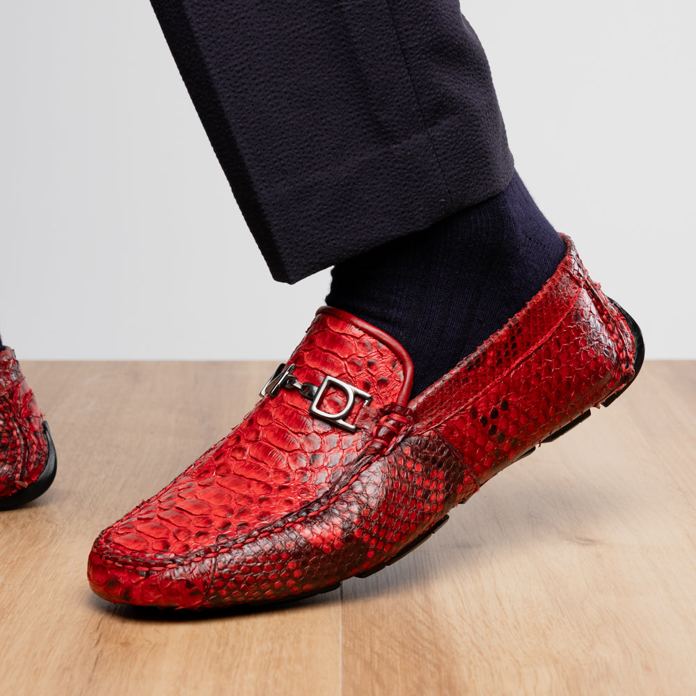 A pair of Pia Red loafers by Marco Di Milano—crafted as luxury men’s driver shoes in a striking red snakeskin pattern, featuring black rubber soles and silver buckle detailing, with one shoe upright and the other displayed on its side—showcase the allure of exotic python-inspired style in a distinguished design.