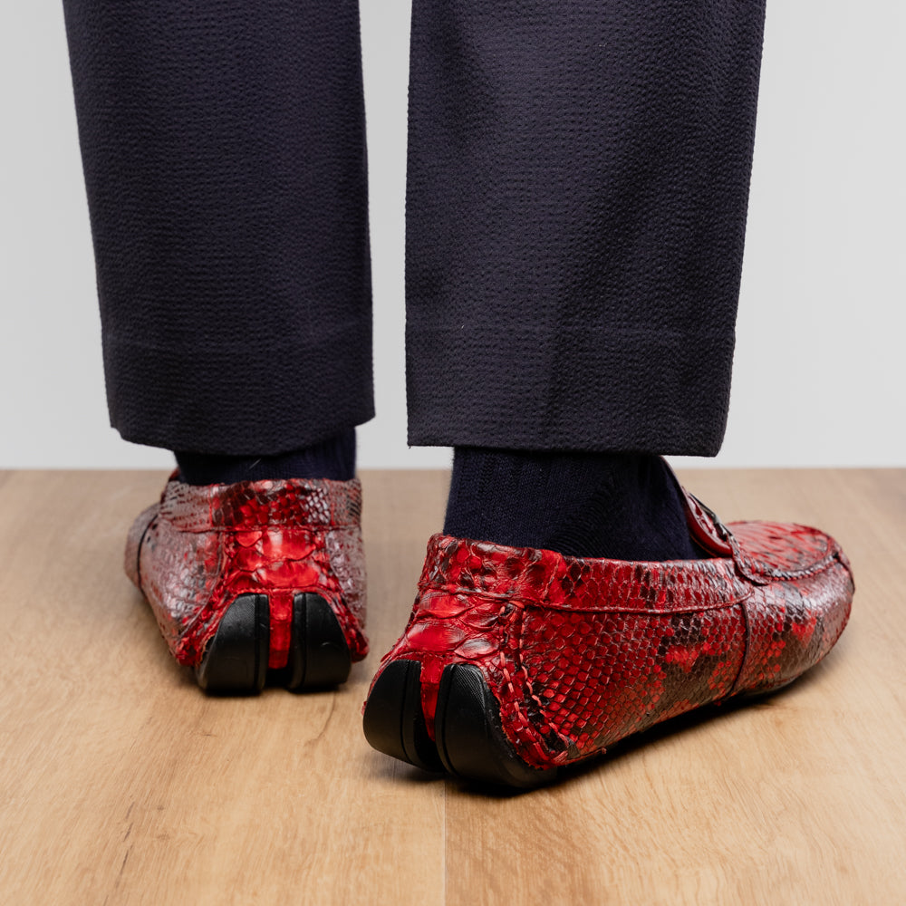 A pair of Pia Red loafers by Marco Di Milano—crafted as luxury men’s driver shoes in a striking red snakeskin pattern, featuring black rubber soles and silver buckle detailing, with one shoe upright and the other displayed on its side—showcase the allure of exotic python-inspired style in a distinguished design.