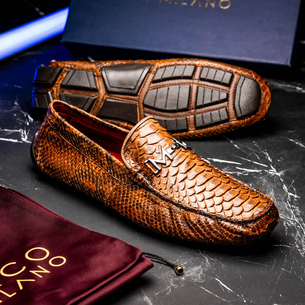 The Marco Di Milano Pia Cognac features brown python skin driver loafers with a metal buckle, visible black rubber soles, and one shoe upright while the other lies flat—a luxury men's shoe designed for style seekers.