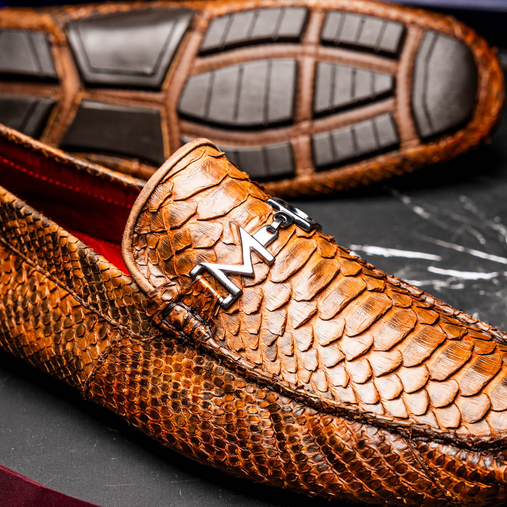 The Marco Di Milano Pia Cognac features brown python skin driver loafers with a metal buckle, visible black rubber soles, and one shoe upright while the other lies flat—a luxury men's shoe designed for style seekers.